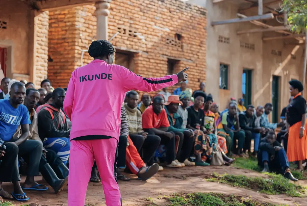 A woman in a pink tracksuit, seen from behind, speaks and points toward a seated adult audience outdoors.