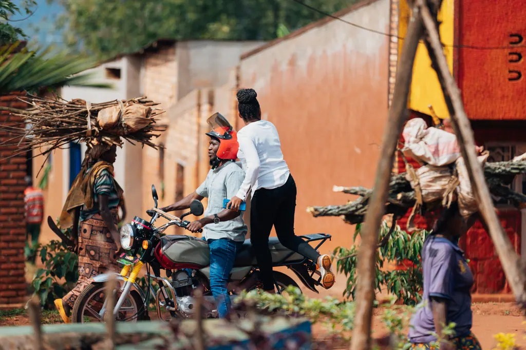 A village street in Kirehe: a woman carries firewood on her head as a motorcycle taxi passes.