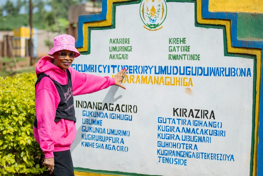 A smiling woman in a pink bucket hat, pink hoodie and a black breast cancer early detection vest points to a painted village sign in Kirehe District, Gatore Sector, Rwantonde Cell, Rwanda.