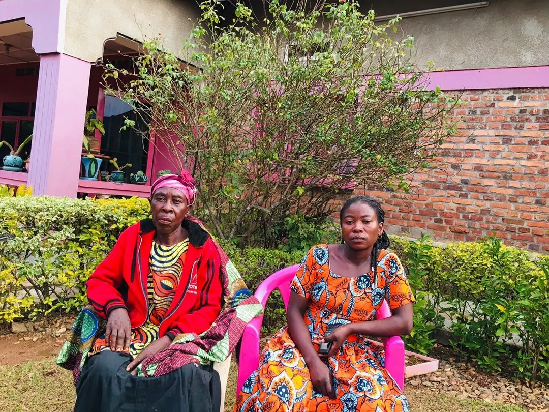 Lea, an older woman in a red jacket and pink headwrap, seated outdoors; a younger woman in an orange dress sits beside her.