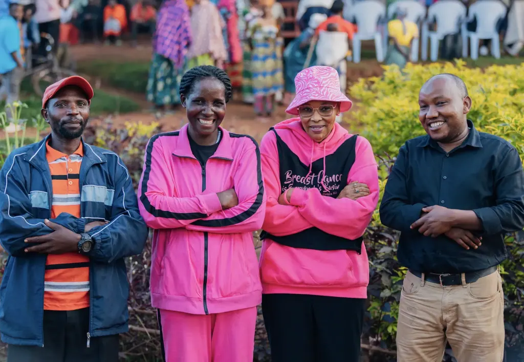 Four smiling members of the Pink Village Movement team; two women wear pink, one in a pink bucket hat and a breast cancer awareness hoodie.